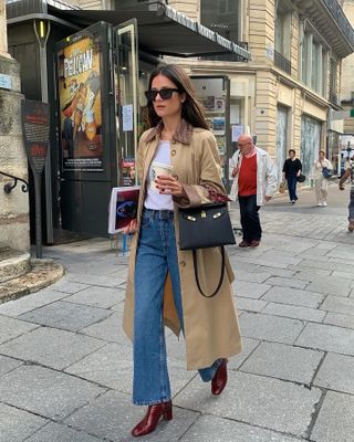 A brunette woman wears a long tan coat with blue jeans and red patent leather boots.