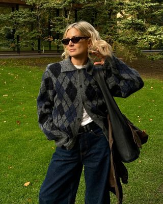 British fashion influencer Lucy Williams poses in Copenhagen wearing an our Legacy argyle cardigan sweater, black oval sunglasses, a white t-shirt, and dark-wash jeans