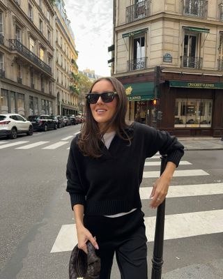 Woman wearing black cashmere sweater, black jeans, Black sunglasses, and Bottega Veneta bag and standing on Paris street.