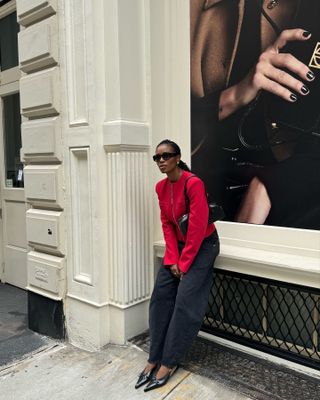 Woman wearing red sweater, black jeans, black slingback kitten heels, and black bag and leaning against SoHo wall.