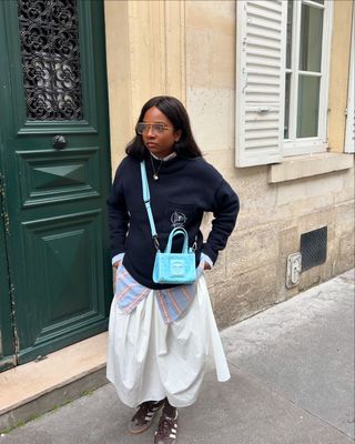 Ryan Norville in a navy sweater, white midi skirt, Adidas sneakers, and a Telfar bag.