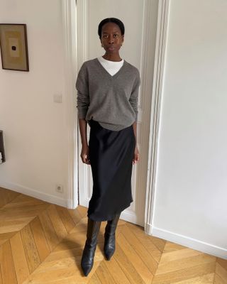 Woman standing in front of white wall, wearing black satin skirt, black boots, white t-shirt and grey sweater.