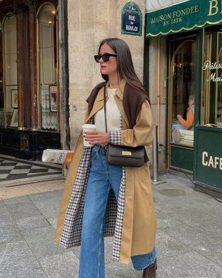 Woman wearing white shirt, blue jeans, brown trench coat and brown leather bag, walking on the streets of Paris.