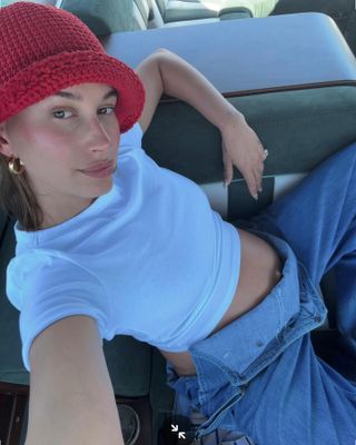 Hailey Bieber wears a red crochet hat, white Joe's Jeans baby tee, and denim overalls.