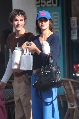 Kaia Gerber wears a blue hat, navy v-neck sweater, blue sweatpants, and a black Balenciaga bag.