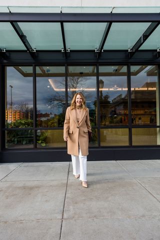 A Nordstrom stylist wearing a long brown coat with a tan sweater and white jeans.