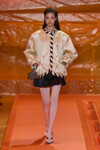 Model at the S/S 24 Louis Vuitton runway show wearing a yellow and black satin padded jacket with a black miniskirt and tights.