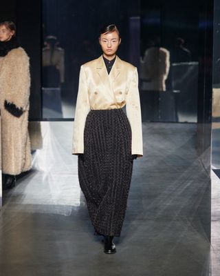 Model at the Eenk F/W 24 runway show wearing a yellow satin blazer with pinstripe barrel-leg pants.