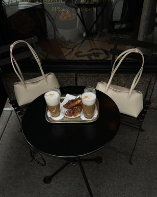 @thecarolinelin and a friend showcase their matching cream handbags, perching them on black cafe seats in front of two foamy coffees