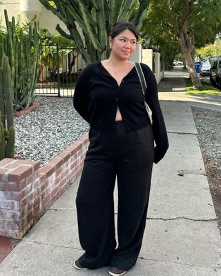 Influencer Marina Torres poses on a sidewalk wearing a black cardigan sweater, black satin pants, and sneakers.