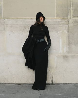 Lena wears a black hat, black long sleeve maxi dress, black belt, and black leather gloves.