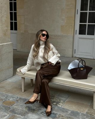 Woman wearing white sweater, brown pants, and leopard shoes while sitting on a bench.