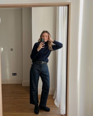 Brittany Bathgate wearing a navy sweater, dark-wash jeans, and black Clarks Wallabee shoes.