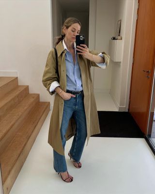 Style influencer Anouk Yve poses for a mirror selfie wearing a trench coat over a blue button-down shirt with a white collar and white cuffs, jeans, and black strappy heeled sandals.