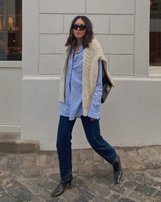 Fashion influencer Debora Rosa walking on a city sidewalk wearing black sunglasses, a blue-button-down shirt, cable-knit sweater over the shoulders, dark-wash straight-leg jeans, and square-toe heeled boots.