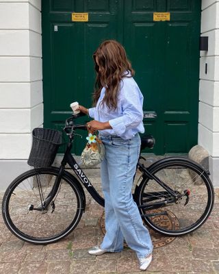 Norwegian fashion influencer Nnenna Echem standing next to an all-black bike in Copenhagen wearing a blue button-down shirt, relaxed jeans, and silver ballet flats.