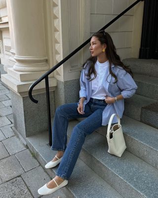 Fashion influencer Felicia Akerstrom sitting on steps outside a building in Stockholm, Sweden wearing an open blue button-down shirt, white T-shirt, straight-leg jeans, and white Mary Jane flats.