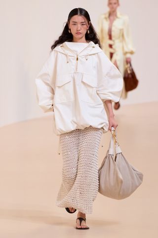 A model at the S/S 25 Zimmermann show wearing a white cotton anorak with sporty details with a crochet maxi dress, sandals, and a big bag.