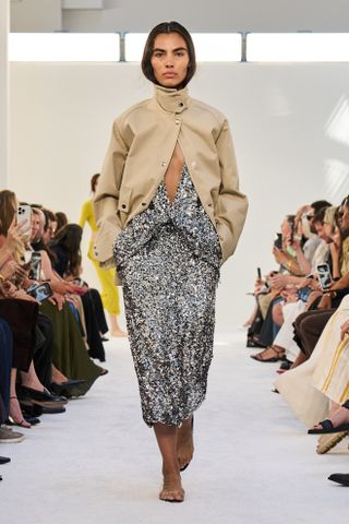 A model at the S/S 25 show at the Brandon Maxwell wearing a khaki funnel-neck utility jacket with a silver sequin low-cut dress and naked flat sandals.