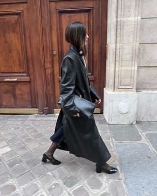 Woman wearing leather trench coat, black shoes, black tights, and black leather clutch standing in front of Paris wooden door.