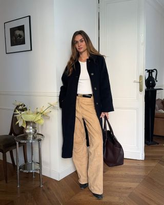 French fashion influencer Anne-Laure Mais posing in her Paris apartment wearing a long black coat, basic white tee, black belt, tan pants, brown tote bag, and black shoes