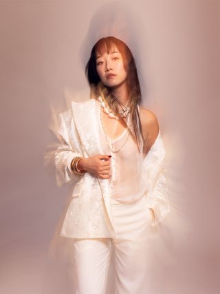 XO, Kitty actress Gia Kim poses wearing a Mariusz Brzezinski white tank top, shirt, and wide-leg trousers with layered necklaces and bracelets.