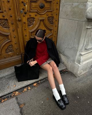 @maryljean wears a red jumper with a black jacket, beige pleated mini skirt, socks and loafers