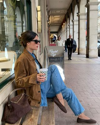 Woman wearing a denim button-down and jeans with a brown suede jacket and a brown suede handbag.