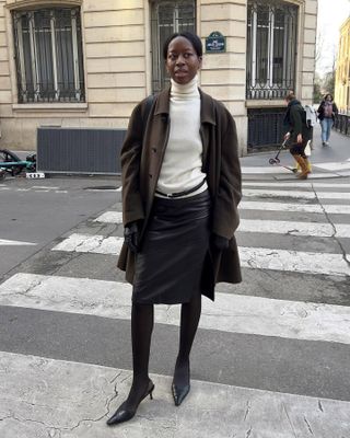 Sylvie wears a brown jacket, ivory turtlneck, leather skirt, black tights, black leather gloves, and slingbacks.