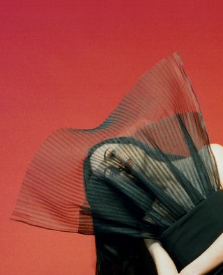 Cazzie David poses in front of a red backdrop wearing a black Nina Ricci dress with sheer veil detailing covering her face.