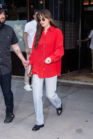 Dakota Johnson wears a red button-down shirt, Levi's jeans, black loafers, and round sunglasses.