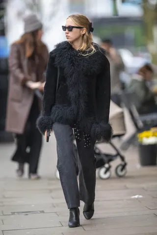 Lila moss wears a black fur penny lane coat, gray jeans, and black leather boots.