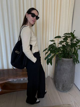 Anna wears black sunglasses, a black liffner bag, black pants, and a cream shirt.