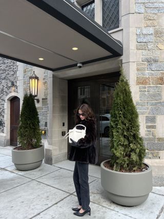 Eliza Huber outside of an apartment building in NYC wearing a brown fur coat, black jeans and shoes, and a cream bag with brunette hair.