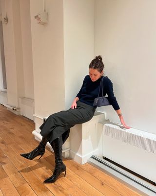 Eliza Huber in the office wearing a navy blue cropped cashmere sweater, charcoal gray midi skirt, navy blue Alaïa bag, and black boots with her brown hair in a bun.