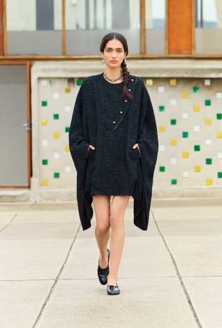 A Chanel model walking in the Resort 2025 show wearing a black cape dress and patent leather moccasins.