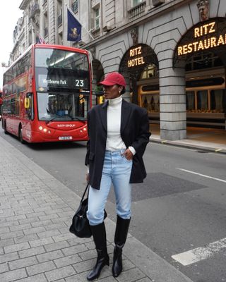 Marilyn wears a red baseball cap, white turtleneck, black blazer blue jeans, black knee-high boots, and a black leather tote bag.