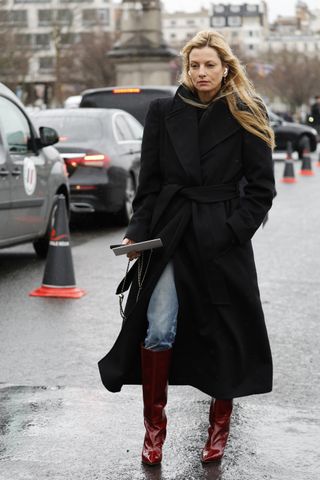 A woman wears a long black coat, blue jeans, and red knee high boots.