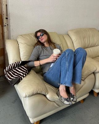 French British fashion influencer Camille Charriere lounges on a sofa wearing oversize cat eye sunglasses, a gray cardigan, zebra-print bag, jeans, and metallic silver Margiela flats.