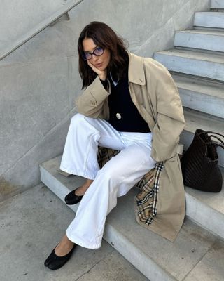 influencer Debora Rosa sitting on cement steps wearing trend eyeglasses, a Burberry trench coat, black top, pendant cord necklace, white relaxed jeans, and black Margiela ballet flats