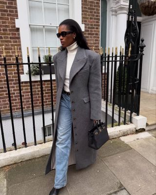 British influencer Marilyn Nwawulor-Kazemaks walking on a London sidewalk wearing black oval sunglasses, a cream turtleneck top, a gray double-breasted coat, belt, Hermes bag, straight-leg jeans, and black boots