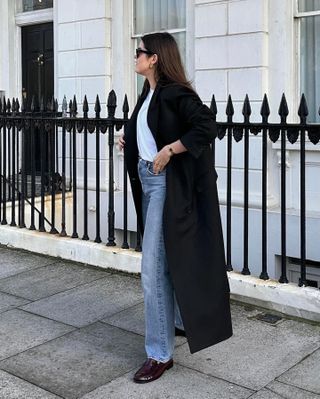 British fashion influencer Anna Newton poses on a London sidewalk wearing a black maxi coat, white tee, straight-leg jeans, and loafers