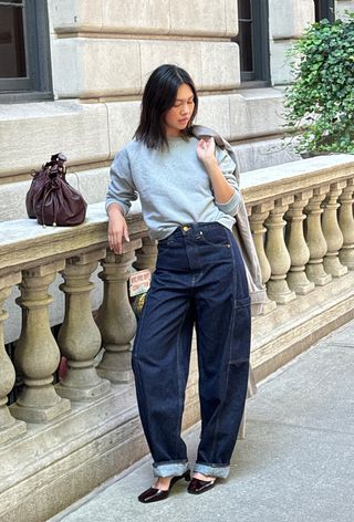 A woman holding a trench coat over her shoulder and wearing a gray sweatshirt styled with dark-wash barrel-leg jeans, brown square-toe kitten heels, and a brown handbag.