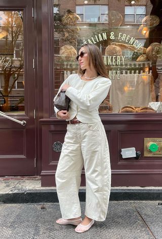 A woman standing in front of a bakery wearing a gold choker necklace, white blazer, brown handbag, white barrel-leg jeans and white mesh ballet flats.