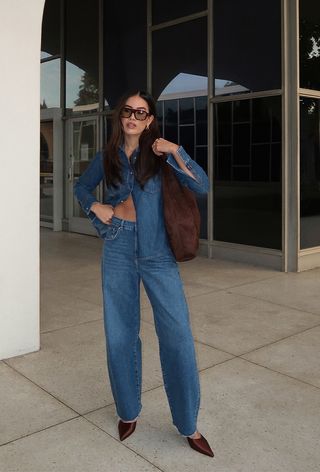 A woman standing next to a glass building wearing a medium-wash denim button-down shirt with matching barrel-leg jeans, brown aviator sunglasses, a brown suede bag, and brown satin heels.