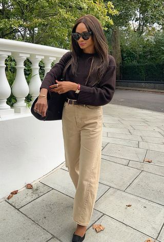 A woman walking down the sidewalk wearing black sunglasses, a brown crewneck sweater, a brown belt, light brown barrel-leg jeans, brown pumps, and a brown tote bag.