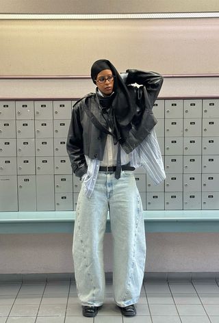 A woman standing inside a mail room wearing a brown leather funnel-neck jacket over a blue striped button-down shirt layered over a white button-down shirt, a black belt, light-wash barrel-leg jeans, black ankle boots, and a black crossbody bag.