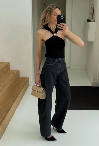 A woman standing in the entryway of her home taking a mirror selfie wearing a black halter top with dark-wash barrel leg jeans, black pointed pumps, and a cream clutch bag.