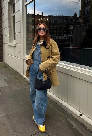 A woman standing outside wearing a barn jacket with a tan sweater, barrel-leg overalls, yellow suede sneakers, and a black shoulder bag.