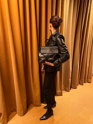 Eliza Huber wearing a leather blazer and black sheer skirt and carrying a black Bottega Veneta Ciao Ciao bag in a room with gold curtains.